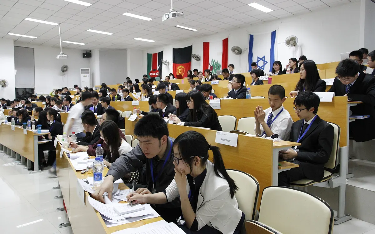 A large committee room at Beijing Model United Nations, with delegates seated in rows of desks under a line of national flags