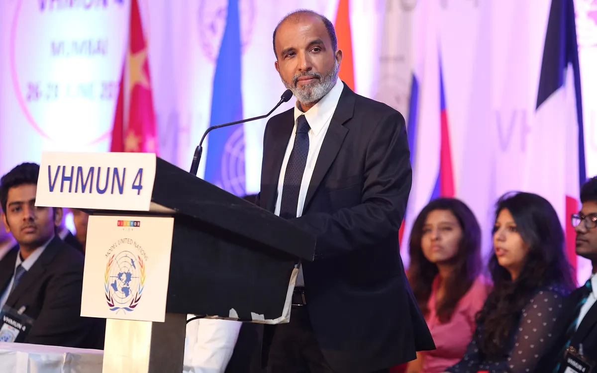 A speaker in a dark suit addressing a Model United Nations conference from a podium, with delegates and flags behind him